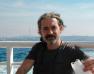 Photo d'un homme sur un bateau qui regarde l'objectif avec un masque chirurgical blanc à la main ainsi qu'un verre en plastique